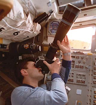 Kaleri with long lens camera, photographs Mir through shuttle flight deck windows