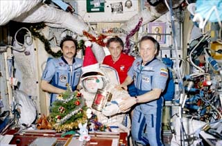 Mir 24 crewmembers posing with a santa doll in a Sokol suit, small tree, decked boughs, and floating presents.